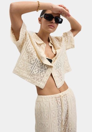 Chemisier court en dentelle beige clair, avec des manches courtes et des motifs décoratifs. Assorti à un pantalon élastique à taille haute.