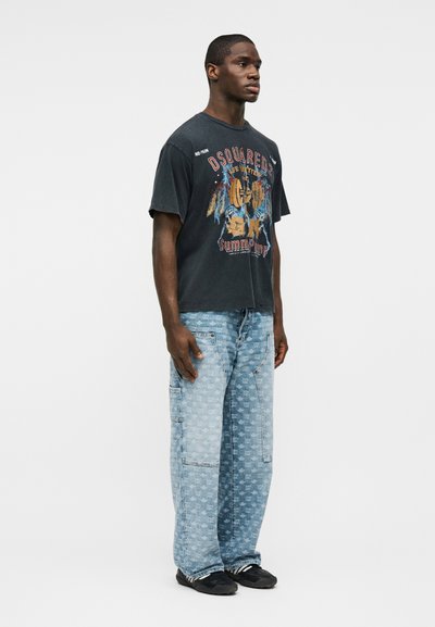 Νεαρός άνδρας σε προφίλ φοράει μαύρο μπλουζάκι με σχέδιο και ανοιχτόχρωμο τζιν cargo με σχέδιο, καθώς και μαύρα αθλητικά παπούτσια.