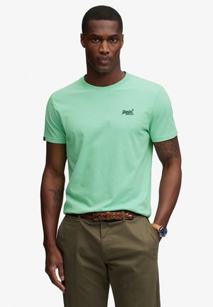 Mann trägt ein hellgrünes Superdry-T-Shirt, olivfarbene Hose, braunen geflochtenen Gürtel und eine Armbanduhr, steht mit einer Hand in der Tasche.