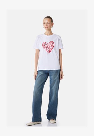 Kvinde iført hvid t-shirt med rødt hjerte og blomsterdesign kombineret med blå jeans og hvide spidse flade sko, stående mod en ensfarvet baggrund.