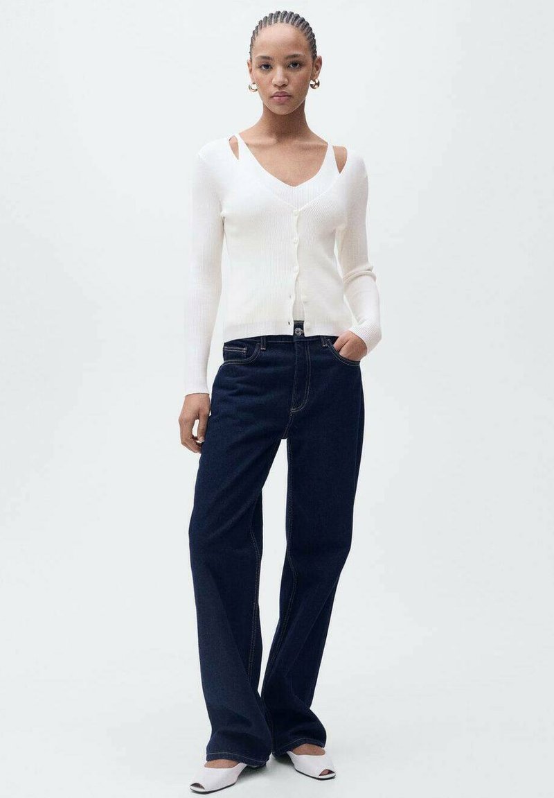 Weißer gerippter langärmeliger Cardigan mit Ausschnitt an den Schultern, kombiniert mit dunkelblauen weiten Jeans und weißen Absatz-Mules.