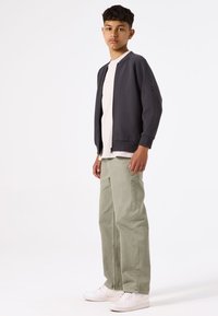 Chaqueta bomber gris oscuro sobre una camiseta crema clara, combinada con pantalones verde claro y zapatillas blancas, de pie frente a un fondo sencillo.