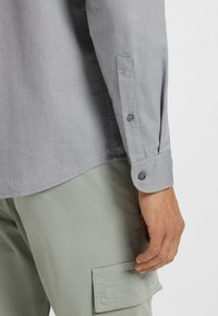 Camisa de manga larga gris con puños abotonados, que presenta una textura suave y un corte recto, combinada con pantalones cargo verdes claros.
