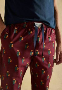 Bordo jogger bikses ar atkārtotu zaļā mēteļa lapsu, kas tur maisiņu, rakstu, valkā ar tumši zilu kreklu, rokas atviegloties pie sāniem.