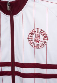 Hvit treningsjakke med røde vertikale striper, med en mørkerød glidelås og brodert logo med "UNFR CREW" og teksten "IKKE KJØR MED OSS."