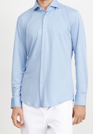 Homme portant une chemise bleu clair à manches longues boutonnée et un pantalon blanc, visible du cou jusqu’aux hanches sur un fond uni.