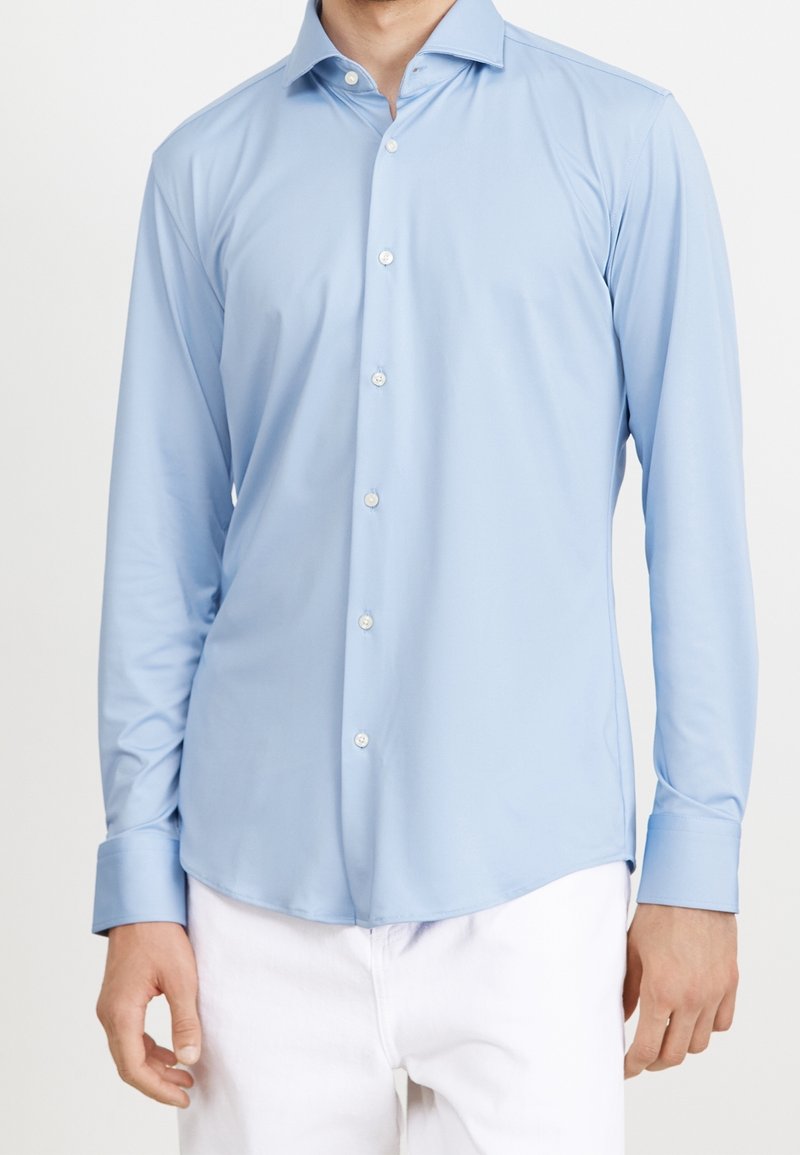 Homme portant une chemise bleu clair à manches longues boutonnée et un pantalon blanc, visible du cou jusqu’aux hanches sur un fond uni.