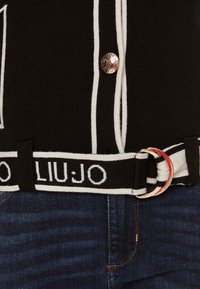 Top in maglia nera con righe bianche, un bottone rotondo in argento e una cintura brandizzata nera e bianca con una fibbia ad anello metallico.