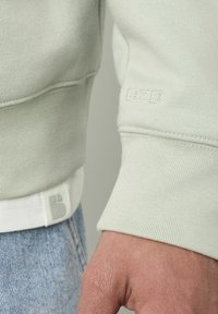 Sweatshirt vert clair avec des poignets côtelés, présentant un logo "BZB" embossé et un ourlet cousu avec une bande blanche contrastante.