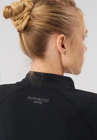 Fekete edzőfelső magas nyakkal, lapos varrással és a hátoldalon fényvisszaverő logóval. Sima textúra, minimalista dizájn.