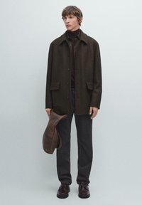 Cappotto di lana marrone con quattro tasche frontali, chiusura con bottoni e colletto, abbinato a pantaloni scuri e scarpe di pelle marrone, mentre tiene un oggetto di pelle marrone.