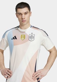 Camisa de futebol Adidas com uma base branca e padrões abstratos em tons pastel, emblema da FIFA e emblema da Espanha. Textura suave e mangas curtas.