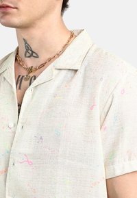 Camicetta a maniche corte di un leggero colore beige, decorata con motivi di spruzzi colorati; realizzata in tessuto testurizzato, con colletto e chiusure a bottone.