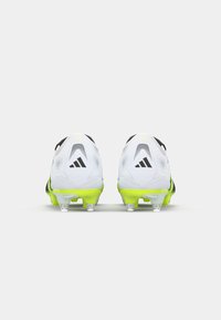 Des chaussures de football blanches et noires avec une semelle jaune, présentant un dessus texturé et des crampons transparents, affichant le logo Adidas de manière proéminente.