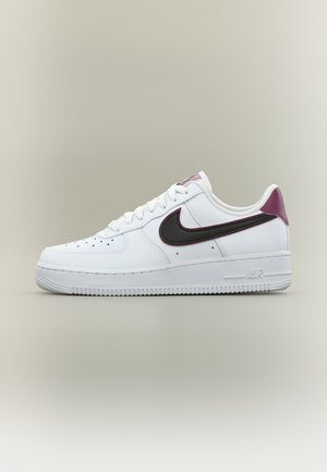 Baskets blanches Nike Air Force 1 avec swoosh noir et bordeaux et languette au talon, vue de côté sur fond beige uni.