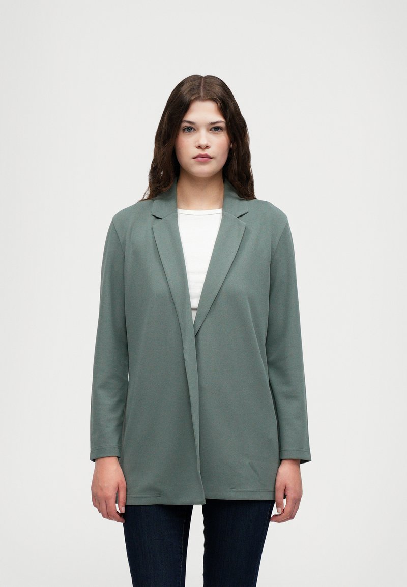 Grønn blazer laget av et mykt stoff, med sjalkrage og åpen frontdesign. Lange ermer og avslappet passform, kombinert med en hvit topp.