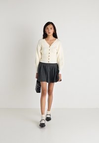 Cardigan cremos cu tricotaj în cable, cu închidere prin nasturi, asortat cu o fustă mini plisată gri. Pantofi negri Mary Jane și o geantă cu buline completează ținuta.