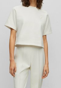 Kobieta ubrana w kremową, krótką bluzę z krótkim rękawem i wysokiej talii spodnie w tym samym kolorze, na szarym tle.