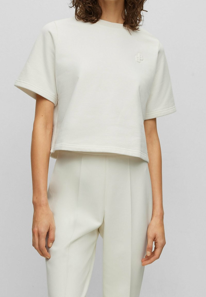 Kobieta ubrana w kremową, krótką bluzę z krótkim rękawem i wysokiej talii spodnie w tym samym kolorze, na szarym tle.