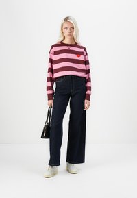 Prugasta jopica v roza in burgundski barvi z grafiko ustnic, v kombinaciji s temnimi širokimi hlačami in belimi supergami, nosi črno torbico.