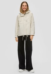 Lichtbeige hoodie-jack met trekkoorden en voorzakken, gecombineerd met zwarte wijdvallende broek en witte sneakers. Gladde textuur, casual ontwerp.