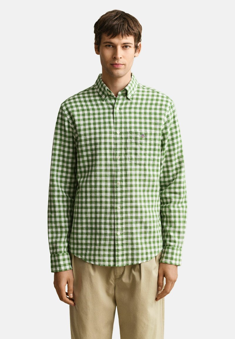 Homme portant une chemise à carreaux verts et blancs boutonnée, avec un pantalon beige, debout devant un fond clair uni.