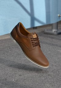 Chaussure en cuir brun avec une texture lisse, dotée d'un design à lacets, d'accents de semelle blanche et d'un petit logo sur le côté.