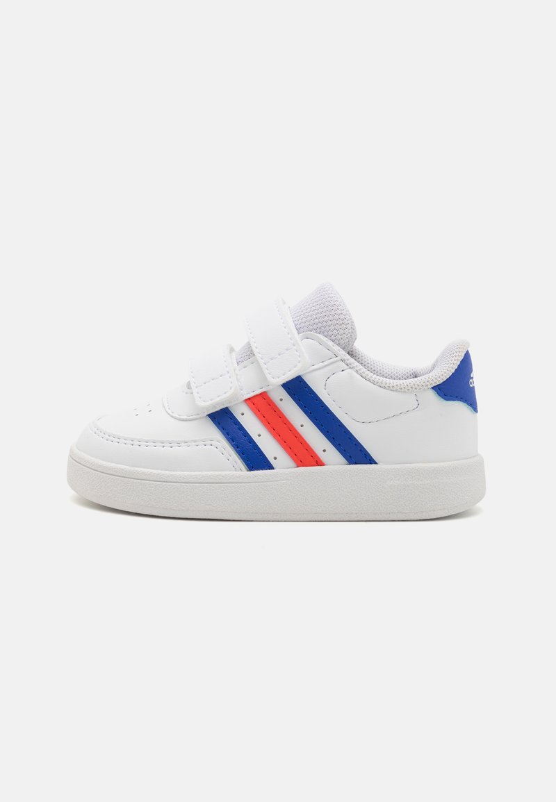 Zapatilla deportiva blanca con rayas azules y rojas, con un upper de cuero suave, dos correas de Velcro y una suela de goma texturizada.