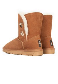 Botas de tobillo de ante marrón con un suave forro de shearling, dos botones decorativos y suelas de goma. Presentan un logo en el lado exterior.