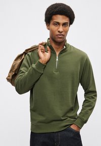 Rohelise lukkudega dressipluus kõrge kaelusega, soonikuga varrukaotste ja äärega. Modell hoiab ühes õlas pruuni kotti. Sile tekstuur.