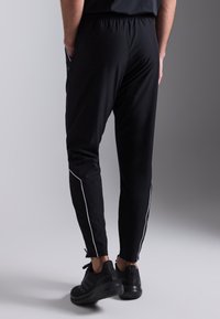 Pantalones deportivos negros con ajuste ceñido, que presentan rayas laterales blancas y una cintura elástica. Incluyen bolsillos y puños ajustables en los tobillos.