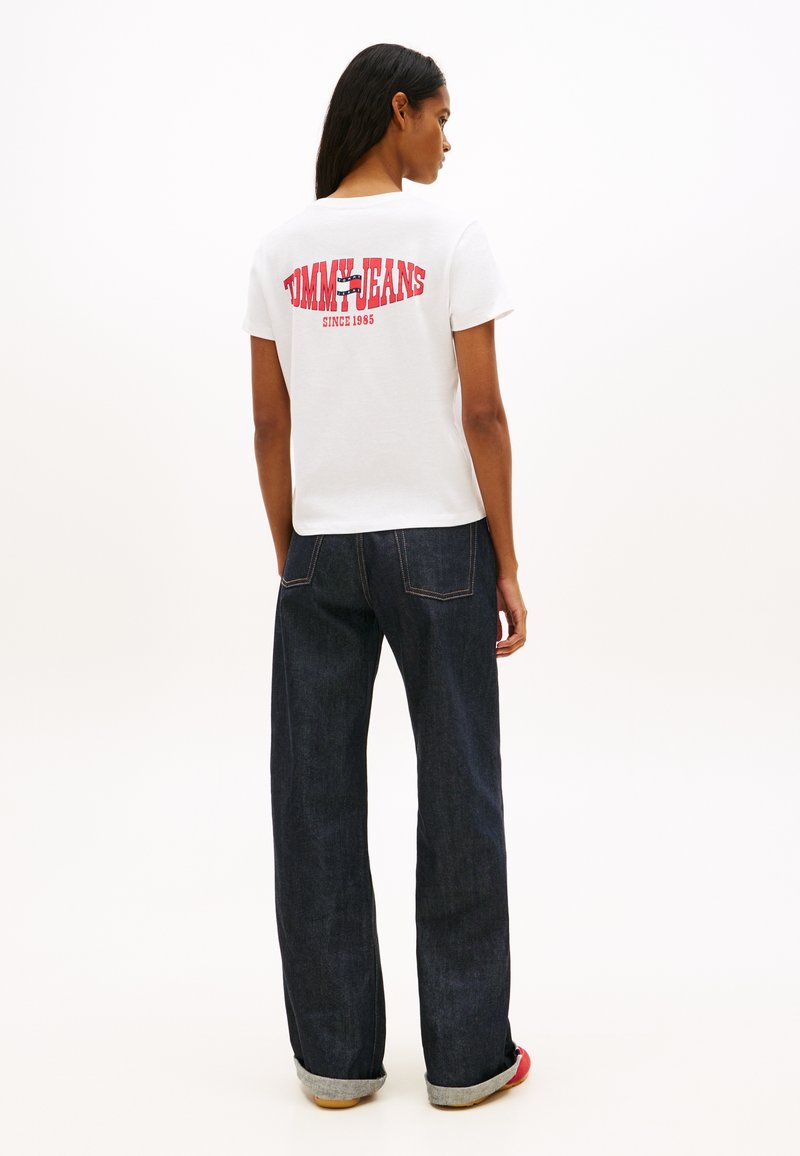 Γυναίκα που φοράει λευκό T-shirt Tommy Jeans με κόκκινο λογότυπο και σκούρο τζιν με πλατιά μπατζάκια, στέκεται γυρισμένη προς τα πίσω μπροστά σε απλό φόντο.
