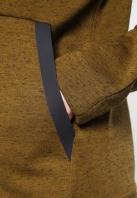 Tela marrón moteada con un ribete gris oscuro. Presenta un bolsillo lateral con costuras visibles en una superficie texturizada.