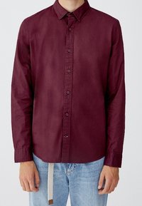 Bordeaux lange mouwen shirt met knoop-onder-nek, voorkant met knoopsluiting en verstelbare manchetten. Combinatie met lichtblauwe jeans met een trekkoord.