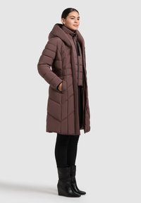 Barna párnázott hosszú kabát kapucnival, cikkcakk mintával, oldalsó zsebekkel, fekete könnyű belső dzseki és fekete leggings fölött viselve.