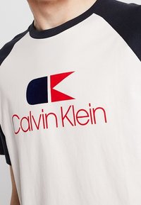 Camiseta Calvin Klein con cuerpo color crema, mangas raglán en azul marino, logotipo audaz en rojo y azul, y una textura de algodón suave.