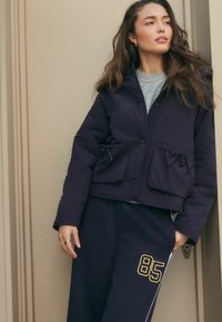Kvinna med lång brunt hår iförd en marinblå vadderad jacka och marinblå sweatpants med gult "85", stående mot en beige vägg.