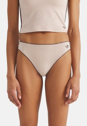 Frau trägt beige High-Cut-Bikinihose und cropped Tanktop mit schwarzem Rand und Adidas-Trefoil-Logos auf beiden Kleidungsstücken.