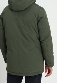 Veste matelassée vert foncé avec une capuche. Dotée d'un tissu lisse et résistant à l'eau avec une coupe droite. Aucun motif visible ni détail matériel apparent.