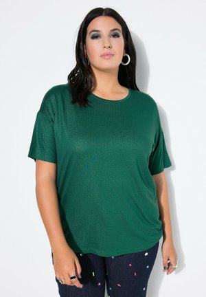 Femme aux cheveux foncés portant un haut vert à manches courtes, un pantalon foncé avec des points de confettis colorés et de grandes boucles d'oreilles, debout devant un fond blanc.