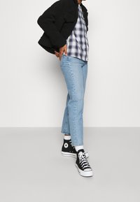 Giacca di felpa nera, camicia a quadri blu e bianca, jeans dritti blu chiaro, sneakers alte nere con suola bianca e calzini bianchi.