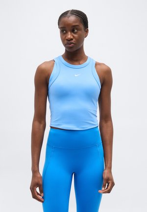 Femme aux cheveux tressés portant un crop top sans manches bleu clair et un legging taille haute bleu vif, debout devant un fond uni.