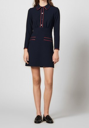 Vrouw in een marineblauwe mini-jurk met lange mouwen, rode biezen, witte knopen en zwarte loafers met kwastjes, staand voor een witte achtergrond.