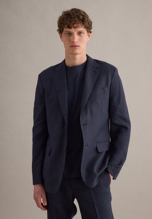 Jonge man met krullend haar die een donkerblauw colbert en bijpassende broek draagt, staande met één hand in de zak tegen een effen achtergrond.