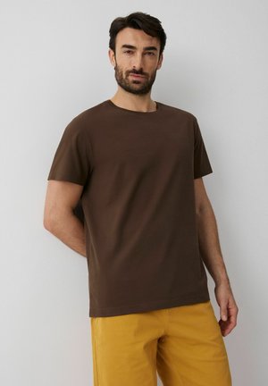 Mężczyzna z ciemnymi włosami i brodą, ubrany w brązowy T-shirt z krótkim rękawem i musztardowe spodnie, stojący przy jasnoszarej ścianie.
