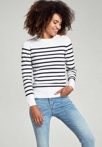 Maglione bianco con righe blu navy, con polsini e colletto a costine, abbinato a jeans azzurri. Il tessuto sembra morbido e aderente.