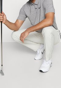 Helles graues Polohemd mit Kragen, schwarzes Logo, weiße Golfhose und weiße Sportschuhe mit schwarzen Akzenten, die einen Golfschläger halten.