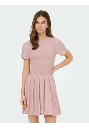 Mlada ženska z dolgimi rjavimi lasmi, oblečena v kratkoročno svetlo roza obleko z teksturo in nabrano krilo, stoji z eno roko na boku.