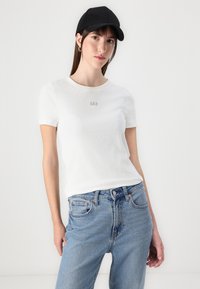 Tricou alb cu mânecă scurtă, cu textură canelată și un mic logo "GAP", asociat cu blugi albaștri cu talie înaltă și o șapcă neagră.
