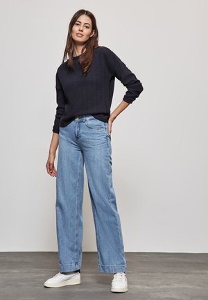 Dunkelblauer strukturierter Strickpullover kombiniert mit hellblauen weit geschnittenen Jeans. Weiße Sneakers vervollständigen das Outfit vor einem neutralen grauen Hintergrund.